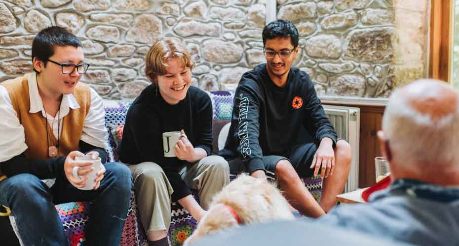 three friends sit on sofa with coffee mugs laughing and petting dog