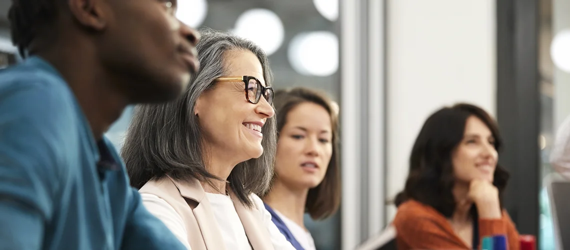 Smiling mature businesswoman sitting with colleagues while looking away. Male and female executives are in board room during meeting.