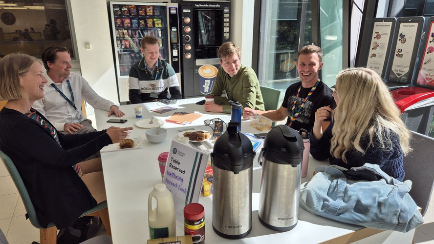 Five adults having a coffee break around a table, smiling and chatting together